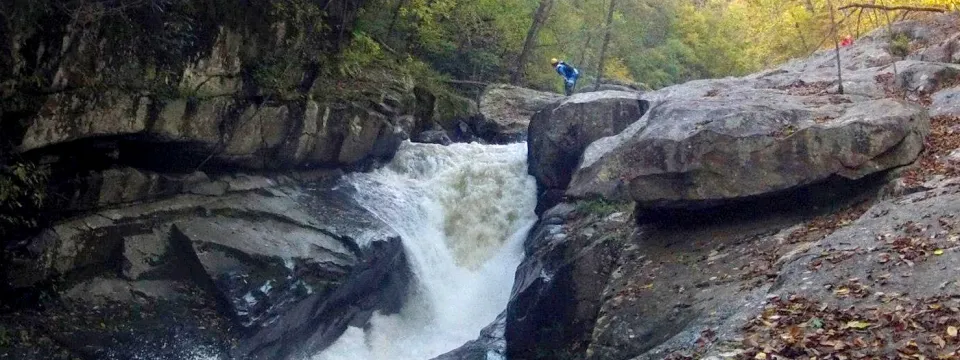 A whitewater gorge: a waterfall pouring between granite slabs, a riverboarder scouting from the rocks above