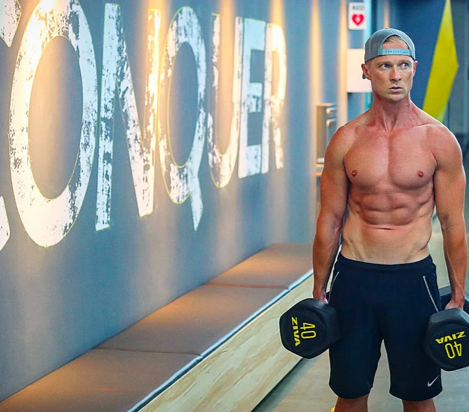 Josh Galt in a gym holding two 40 kg dumbbells under a CONQUER wall graphic